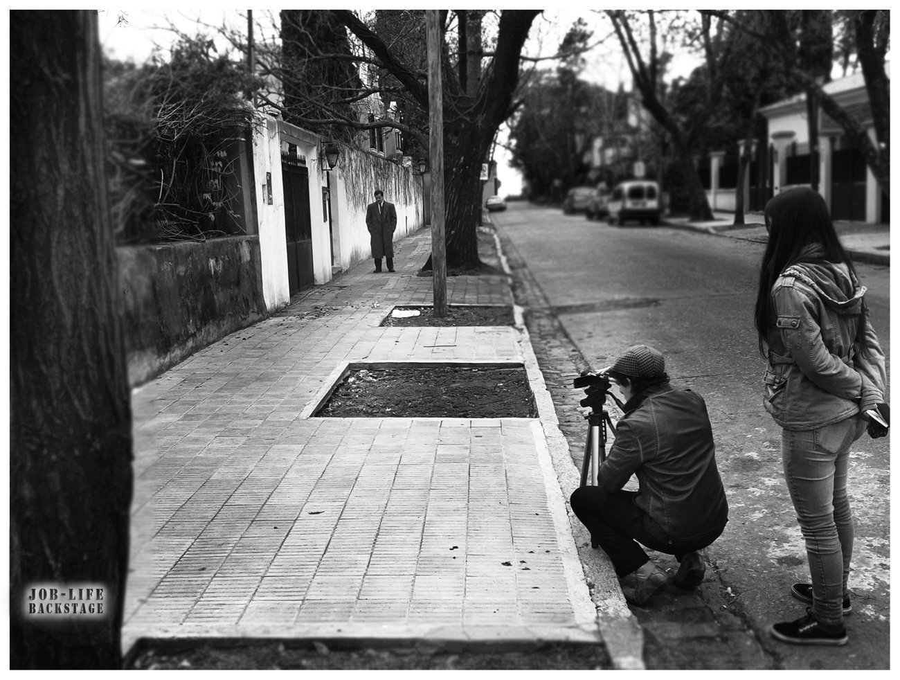 Black and white photograph of a street film shoot: a crouching camera operator films an actor in a long coat on a tree-lined pavement while a crew member watches.