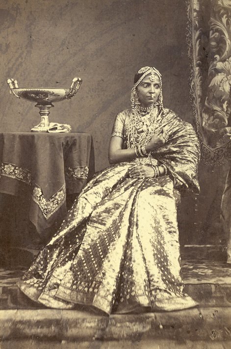Sepia studio portrait of a seated girl in a ceremonial silk sari and jewelled head-dress.
