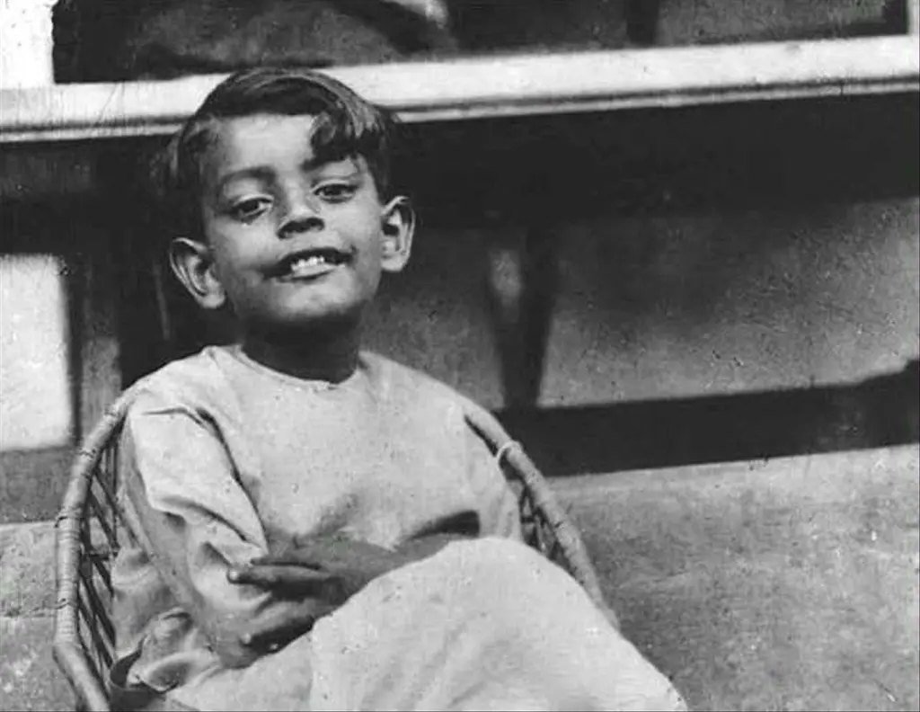 Black and white photograph of the young Satyajit Ray, smiling, seated in a cane chair on a step.