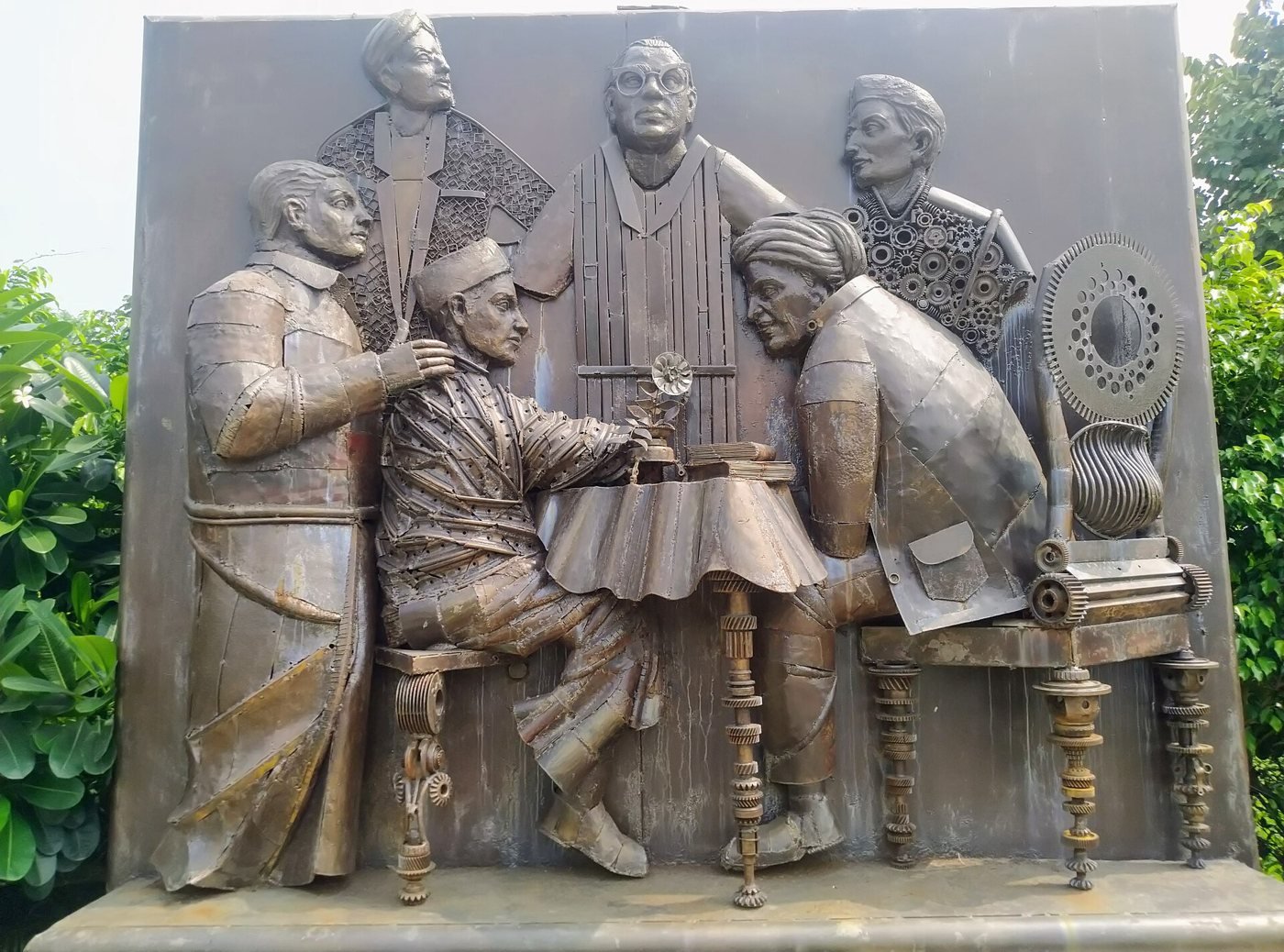 A large metal relief sculpture of six men in early twentieth-century dress gathered around a table, built from welded scrap: gears, sheet metal and machine parts.